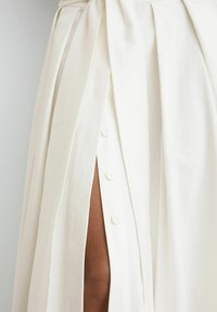 Witte geplooide rok met knopen aan de voorkant en een hoge split die een been onthult tegen een effen lichte achtergrond.