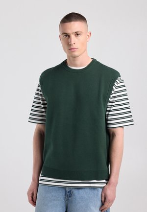 Jonge man met kort haar die een donker groene mouwloze trui draagt over een zwart-wit gestreept T-shirt en lichtblauwe spijkerbroek.