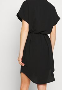 Vestido negro de manga corta, hecho de una tela ligera. Presenta una cintura ceñida con cinturón y un dobladillo asimétrico con una textura suave.