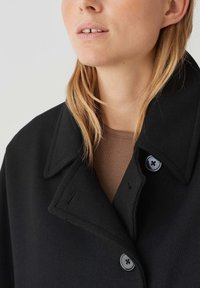 Cappotto intrecciato nero con un ampio colletto, dotato di due bottoni neri. Texture morbida e vestibilità rilassata, indossato su una maglia marrone. Inquadratura ravvicinata.
