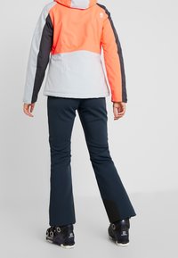 Wasserdichte Ski-Jacke in Orange, Grau und Schwarz mit Kapuze; kombiniert mit dunkelblauen Skihosen und schwarzen Skischuhen mit mehrfarbigen Beschlägen.