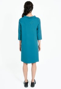 Teal skiftklänning med krage, tre-kvarts ärmar och rak fåll, kombinerad med svarta klumpiga skor. Enkelt, slätt tyg med mjuk textur.