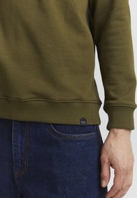 Olivengrønn hettegenser med ribbestrikkede mansjetter og hem, teksturert stoff, med et sekskantet svart merke med "CASUAL" preget. Kombinert med mørkeblå jeans.