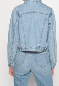 Světle modrá džínsová bunda s přiléhavým střihem, viditelným šitím a kovovými komponenty v pase. Obsahuje klasický límec a zadní yoke.