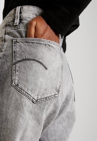 Šedé džíny z denimu s mírně vybledlým povrchem, s zadní kapsou a zakřiveným ozdobným stehem. Ruka je viditelná v kapse.