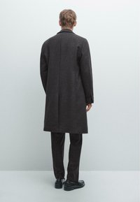 Uomo che indossa un lungo cappotto grigio scuro, pantaloni scuri e scarpe nere, in piedi di fronte a una parete bianca semplice.
