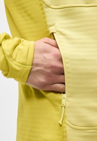 Χέρι εν μέρει μέσα σε τσέπη με φερμουάρ ενός κίτρινου μακρυμάνικου ενδύματος, που δείχνει λεπτομερείς ραφές και υφή υφάσματος.