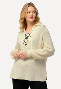 Maglione lavorato a maglia color crema con vestibilità rilassata, dotato di cappuccio, chiusura con lacci sulla parte anteriore e polsini a coste. Fessure laterali evidenti all'orlo.