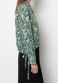 Kobieta w jasnozielonej bluzce z długim rękawem w ciemnozielony nieregularny wzór kropek i czarnej spódnicy, stojąca z boku na białym tle.