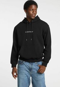 Felpa nera con cappuccio, tasca frontale, coulisse e logo bianco "GUESS JEANS". Indossata con jeans azzurri.