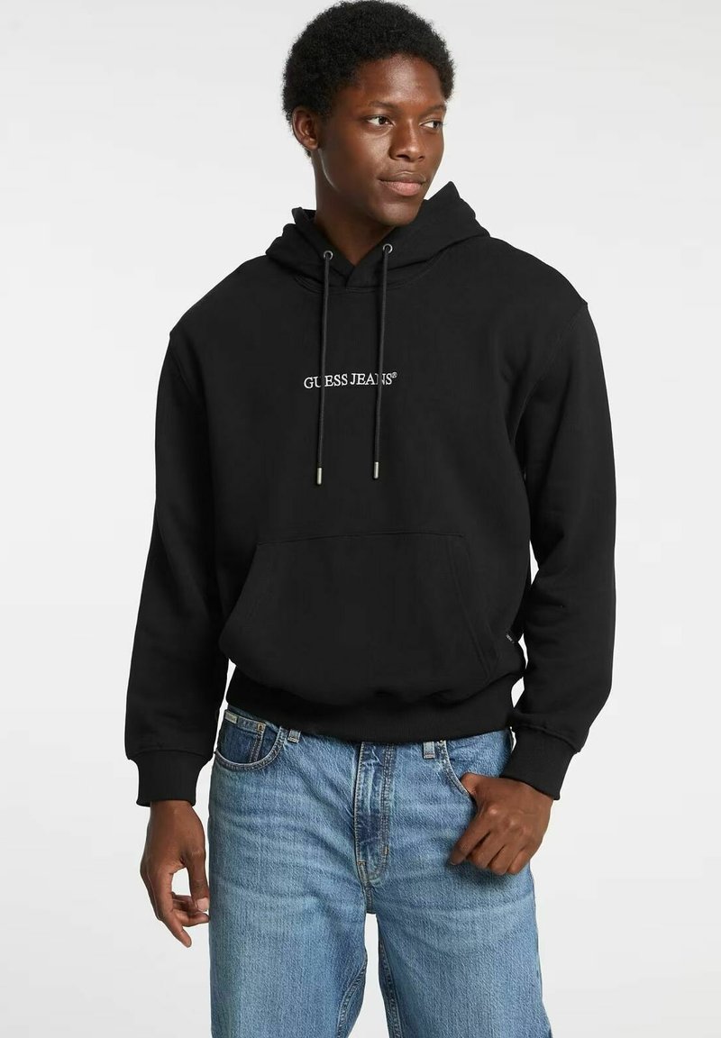 Felpa nera con cappuccio, tasca frontale, coulisse e logo bianco "GUESS JEANS". Indossata con jeans azzurri.