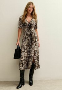Vestido de leopardo en beige y negro, con mangas cortas, de longitud midi y con una abertura, combinado con botas negras altas hasta la rodilla y un bolso negro.