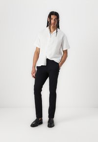 Chemise blanche en lin à manches courtes avec boutons sur le devant, associée à un pantalon noir ajusté et des chaussures noires, sur fond blanc uni.