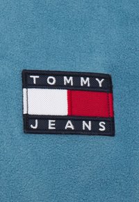 Patch Tommy Jeans sur un tissu doux bleu clair, avec une bordure marine, des rayures horizontales blanches, rouges et bleues, et du texte en lettres blanches.
