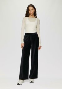 Crème lange mouwen top met een ronde hals, gecombineerd met brede zwarte fluwelen broek. Model draagt witte sneakers.
