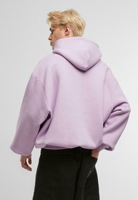 Felpa oversize viola chiaro con cappuccio regolabile, maniche ampie e texture morbida. Indossata con pantaloni cargo neri, che mostrano una vestibilità rilassata.