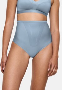 Kvinna som bär ljusblå high-waisted formande trosor och matchande bralette, stående mot en helt vit bakgrund.