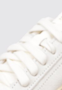 Zapatilla blanca con material de cuero suave, punta redonda, cordones gruesos y sutiles detalles de costura a lo largo de los lados.