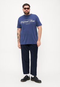 Navy blue cotton t-shirt with white text, paired with dark corduroy pants and black shoes. Accessories include black sunglasses and tattoos.