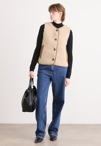 Μπεζ φλις γιλέκο με στρογγυλή λαιμόκοψη, κλείσιμο με κουμπιά και καφέ πλαϊνές τσέπες, φορεμένο πάνω από μαύρη μπλούζα και συνδυασμένο με μπλε τζιν και μαύρη τσάντα.
