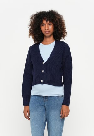 Marineblå kort cardigan med dyp V-hals, ribbestruktur og tre dekorative knapper. Brukt over en lys blå skjorte og jeans.