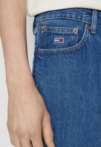 Blå jeans i denim med kopparnitar, synliga sömmar och en liten röd, vit och blå logotypmärke på bakfickan.