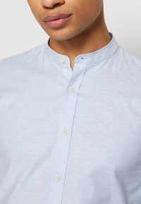 Homme portant une chemise bleu clair boutonnée avec un col mandarin, montrant les lèvres, le menton et la zone de la poitrine sur un fond uni.