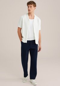 Balta īsām piedurknēm krekls ar pogām, uzvilkts virs baltās topiņa. Tumši zili brīva stila bikses un baltas skriešanas apavus ar akcentu svītrām.