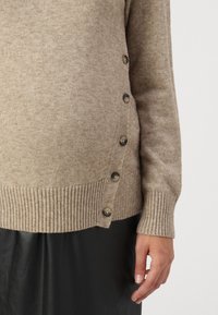 Beige neulepusero, jossa on ribattu helma ja napit sivuilla. Sileä tekstuuri, pitkät hihat ja pehmeä viimeistely.