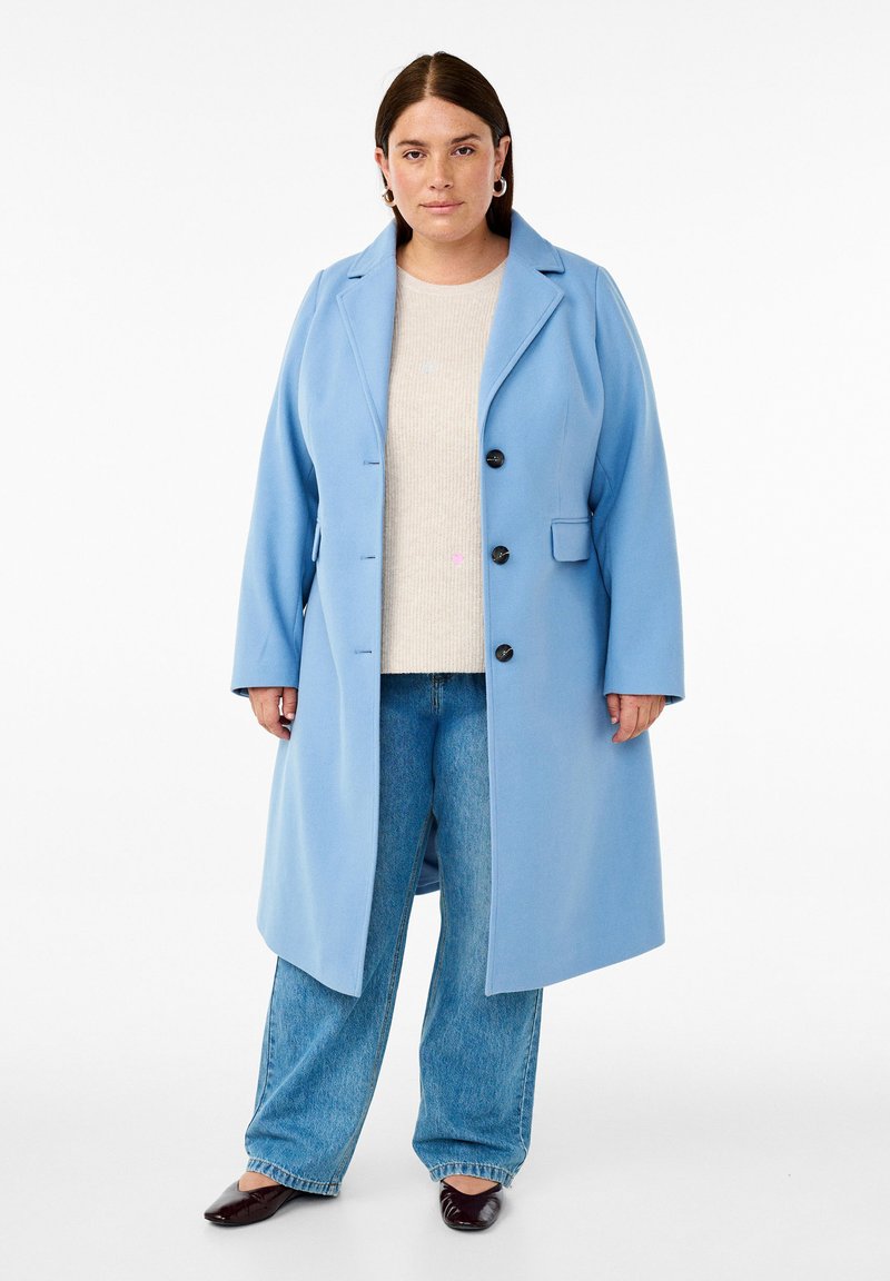 Svetlo modri plašč iz volnene mešanice z izrezanim ovratnikom, enojno zaponko, tremi gumbi in stranskimi žepi, oblečen preko bež puloverja in kavbojk.