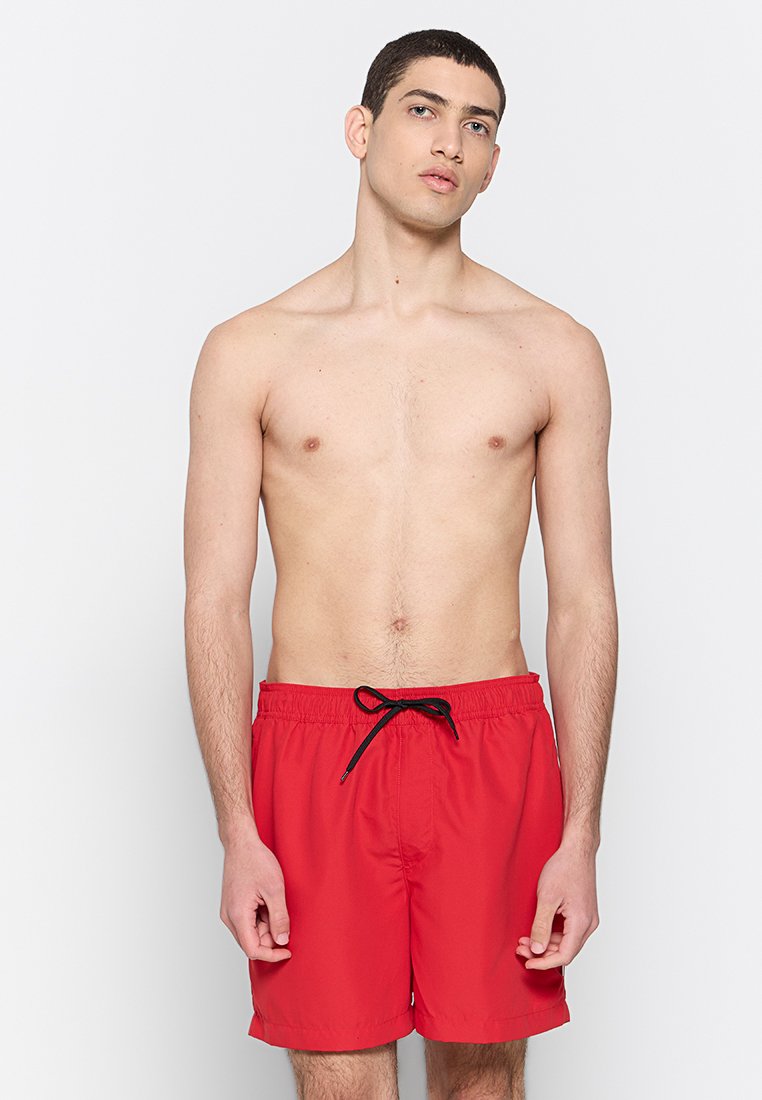 Jeune homme debout devant un fond uni, portant un short de bain rouge avec un cordon noir, regardant légèrement vers le haut.