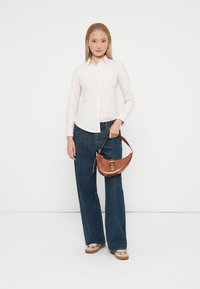Svetloroza srajca na gumbe, modri široki jeans in rjava usnjena torbica z teksturirano površino, ki jo držimo v roki.
