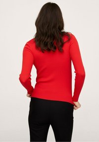 Rode geribbelde lange mouw top, aangesloten ontwerp met een ronde halslijn en textuurstof. Geshowd met zwarte broeken, gezien vanaf de achterkant.