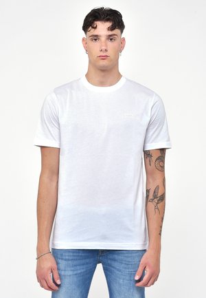 T-shirt bianco in cotone con maniche corte e scollatura rotonda, caratterizzato da un logo sottile sul petto e una vestibilità regolare. Jeans e tatuaggi visibili.