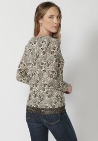 Langärmliges Hemd in Creme mit schwarzem Blumenmuster und Spitzenbesatz am Saum. Weicher Stoff und taillierte Passform, kombiniert mit blauen Jeans.