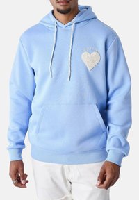 Felpa blu chiaro in tessuto morbido con tasca a marsupio frontale, presenta una patch a forma di cuore testurizzata sul petto e un cappuccio con cordino.