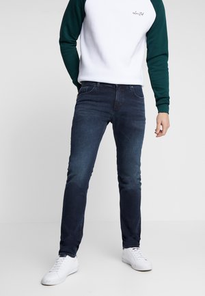 Mężczyzna w dopasowanych ciemnoniebieskich dżinsach, białych sportowych butach i białej bluzie z zielonymi rękawami, stojący na jednolitym jasnym tle.