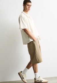 Kortærmet, cremet skjorte med knapper og et tekstureret mønster, parret med tan knælængde shorts og cremede sneakers med brune accenter.