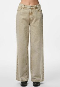 Jeans beige a gamba larga indossati con scarpe nere, con tasche frontali e chiusura con bottone in vita.