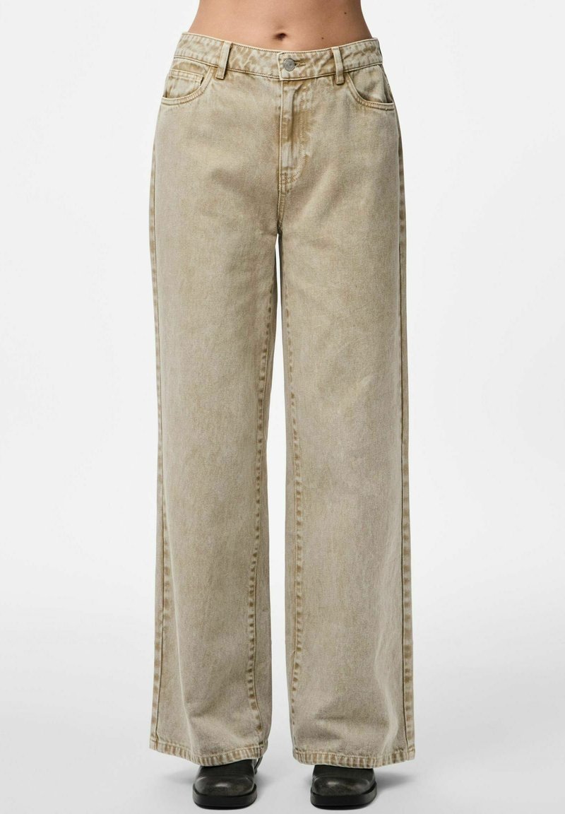 Jeans beige de pierna ancha combinados con zapatos negros, con bolsillos frontales y un cierre de botón en la cintura.