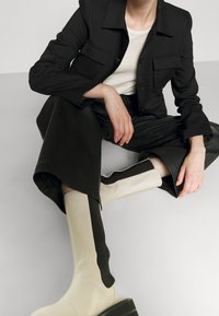 Černý přizpůsobený blazer se dvěma předními kapsami, zkombinovaný s černými kalhotami vysokého pasu a krémovými kozačkami po kolena s kontrastním hnědým pruhem.