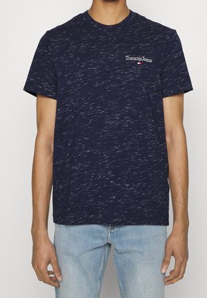 Mand iført en marineblå, plettet Tommy Jeans t-shirt og lyseblå jeans, stående mod en ensfarvet baggrund.