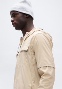 Νεαρός άνδρας με μπεζ μπουφάν με κουκούλα και ανοιχτό γκρι σκουφάκι που κοιτάζει αριστερά, με λευκό απλό φόντο.