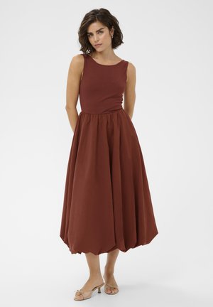 Femme aux cheveux courts et bouclés portant une robe midi marron sans manches avec une jupe volumineuse et talons ouverts beiges, debout devant un fond blanc.