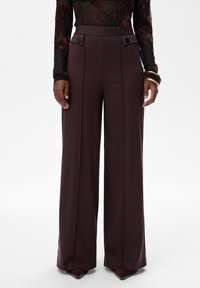 Hoge taille brede bruine broek met voorste knoopflapzakken, gedragen met een doorschijnend bloemen lange mouw top en armbanden om de rechterpols.