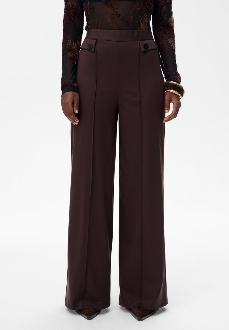 Hoge taille brede bruine broek met voorste knoopflapzakken, gedragen met een doorschijnend bloemen lange mouw top en armbanden om de rechterpols.