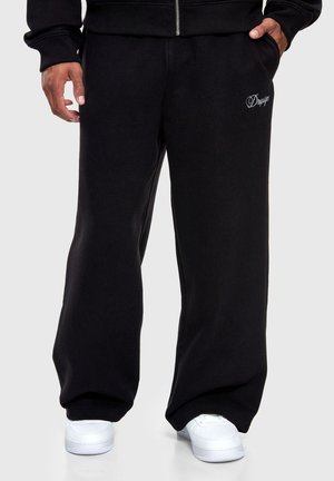 Personne portant un pantalon de survêtement noir à jambes larges avec une broderie blanche sur la cuisse et des baskets blanches, une main dans la poche et l'autre détendue.