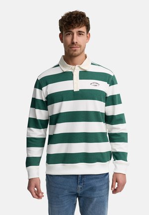 Młody mężczyzna ubrany w zielono-białą koszulkę polo z długim rękawem o szerokich paskach oraz niebieskie dżinsy, stojący na jednolitym tle.