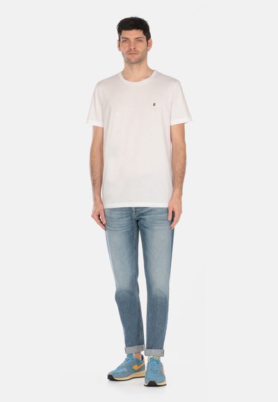 T-shirt bianca in cotone con scollatura rotonda e maniche corte, abbinata a jeans azzurri e sneakers blu con accenti gialli.