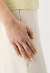 Arany gyűrű egy kerek, tiszta drágakővel, amely egy kezen látható, amely egy textúrázott, redőzött, világos bézs anyagon nyugszik. Rövid, semleges színű körmök láthatók.